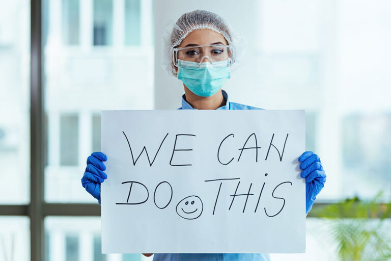 A nurse holding up a sign that reads "We Can Do This" with a smiley face drawn at the bottom.